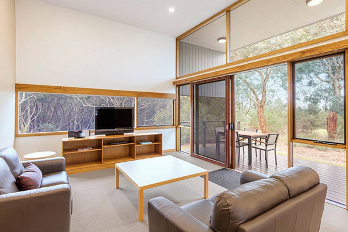 A modern living room with a flat-screen TV, wooden furniture, and large windows offering a view of trees.