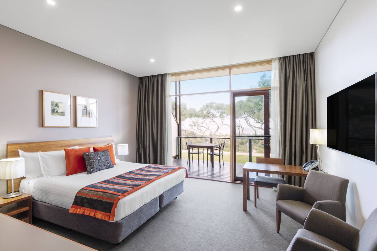 A hotel room with a large bed, a desk, a chair, and a balcony with a table and chairs.
