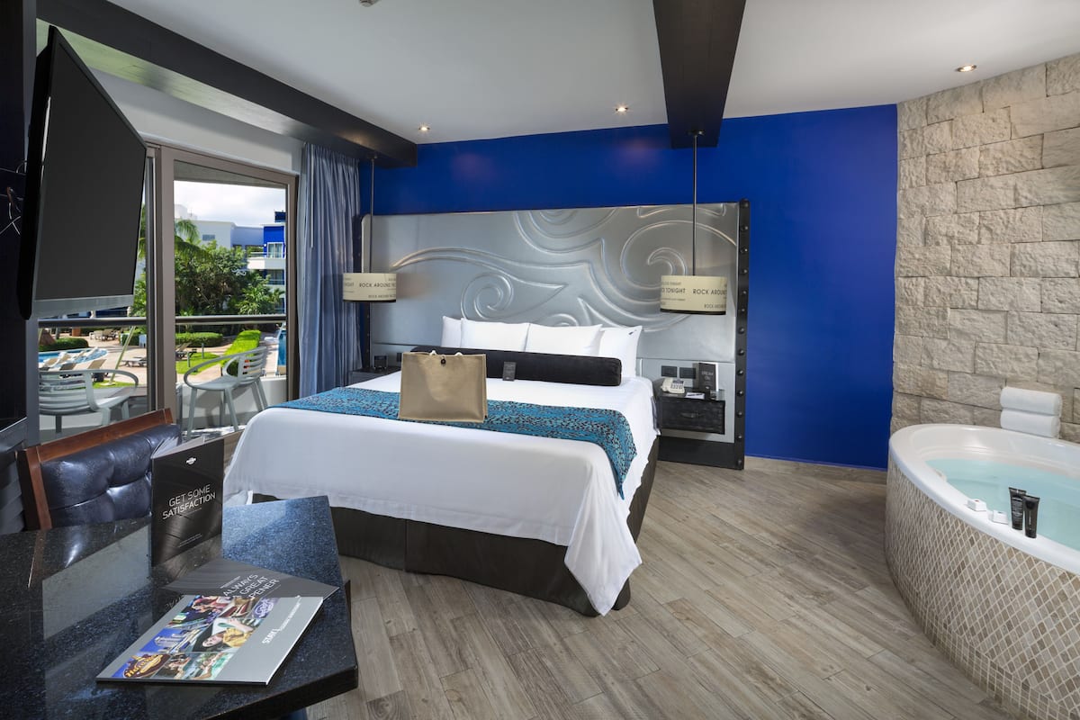 A modern hotel room with a large bed, a sitting area with a table, a bathtub, and a view of the outdoors.