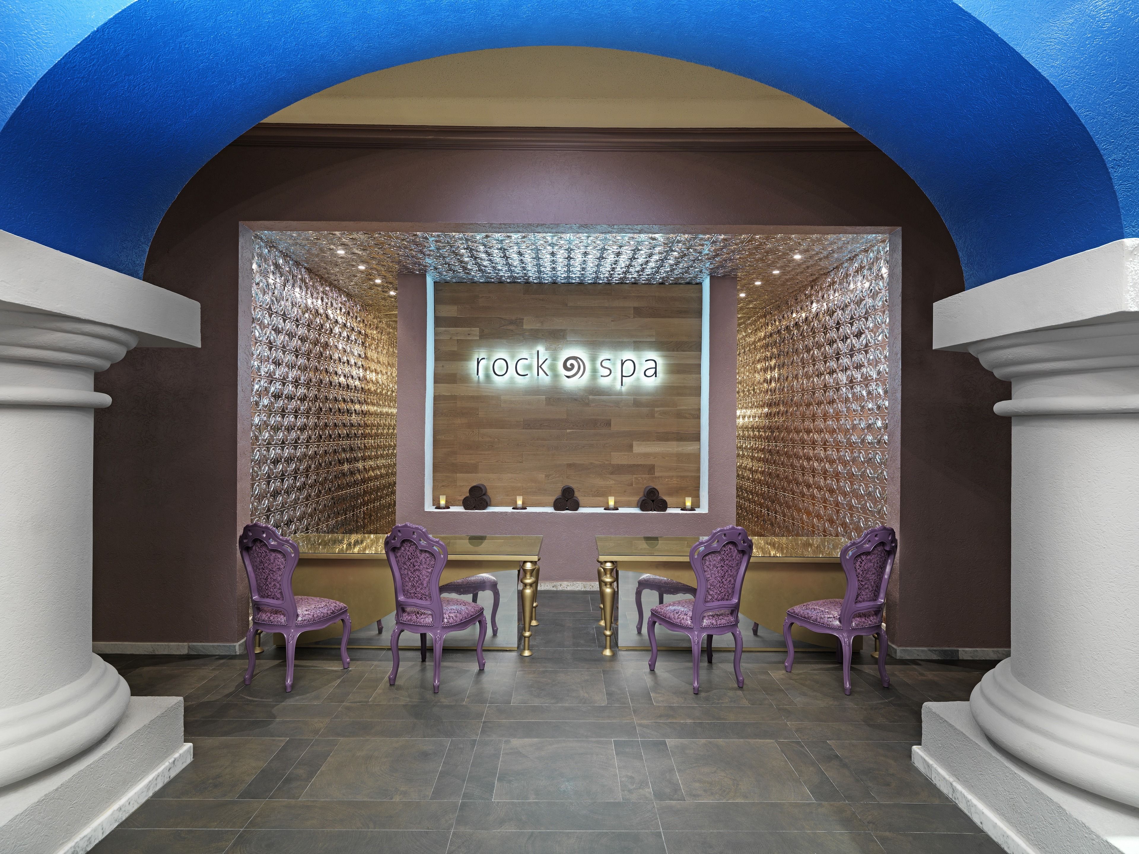 Un espace d’accueil de spa avec une enseigne indiquant « spa aux pierres », deux fauteuils violets et un mur recouvert de miroirs à motifs.