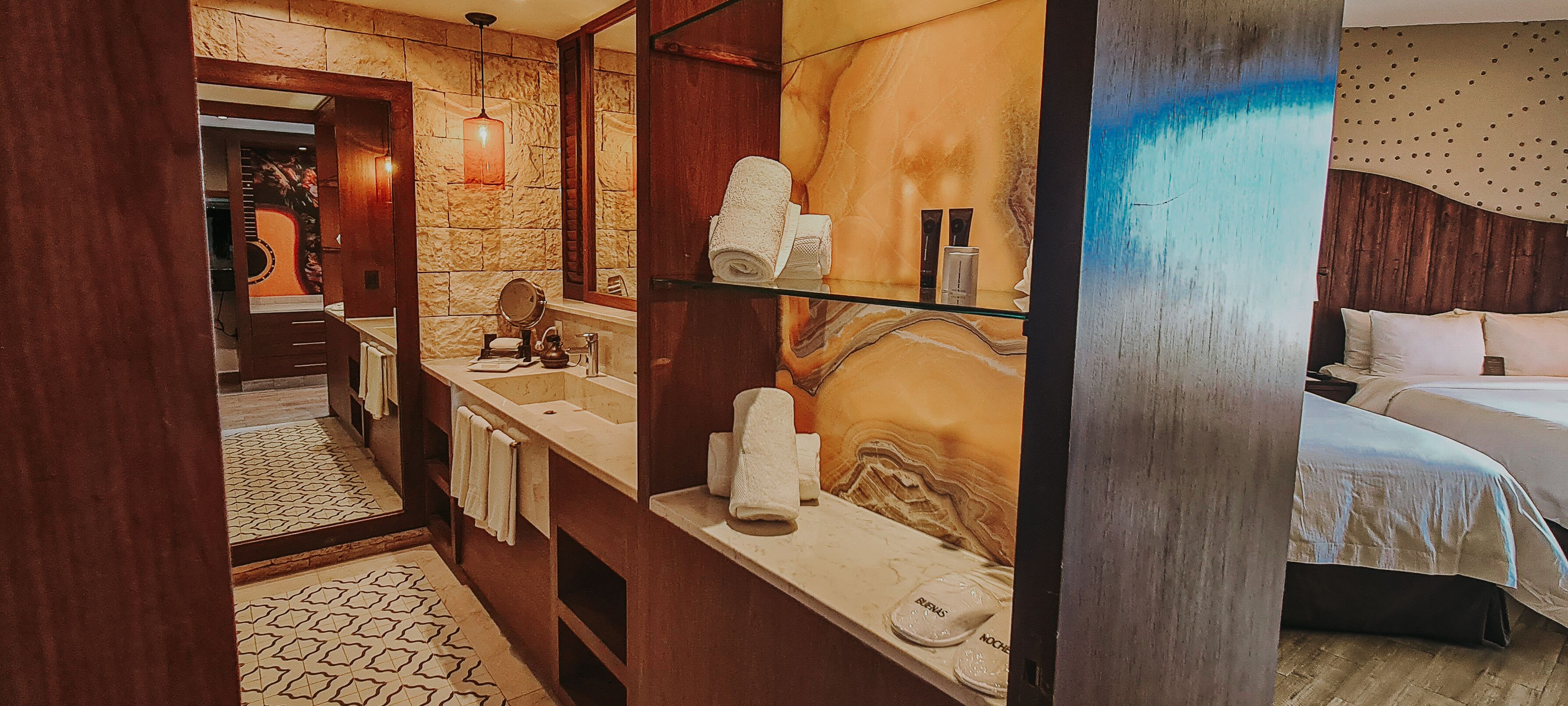 A hotel bathroom with a marble wall, a large mirror, a sink, and a bed with white linens.