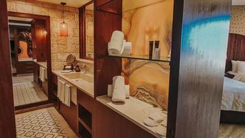 A hotel bathroom with a marble wall, a large mirror, a sink, and a bed with white linens.