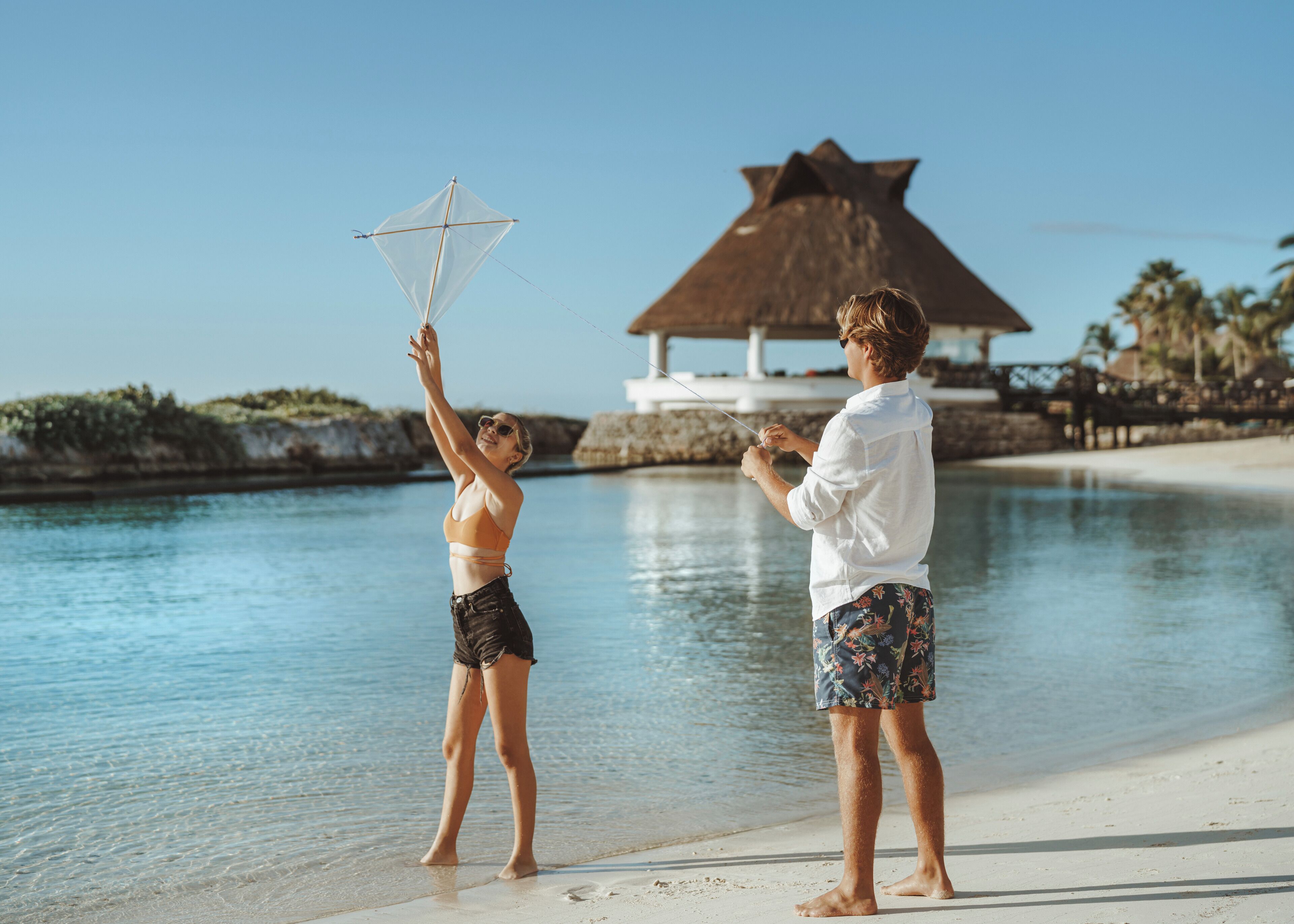 Dos personas volando una cometa en la playa, con una estructura de techo de paja al fondo.