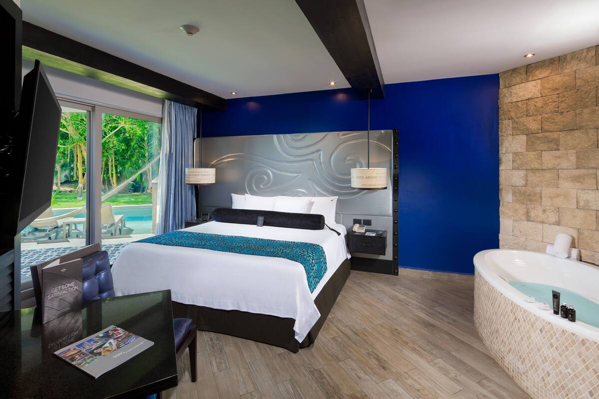 A modern hotel room with a large bed, a separate bathroom with a jacuzzi, and a view of the pool area through the glass door.