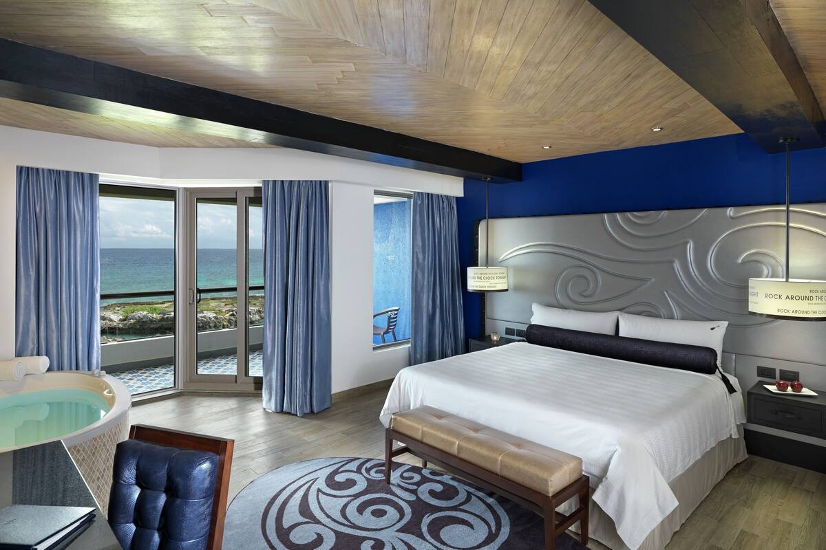 A hotel room with a large bed, a bench, a desk, and a view of the ocean through a sliding door.