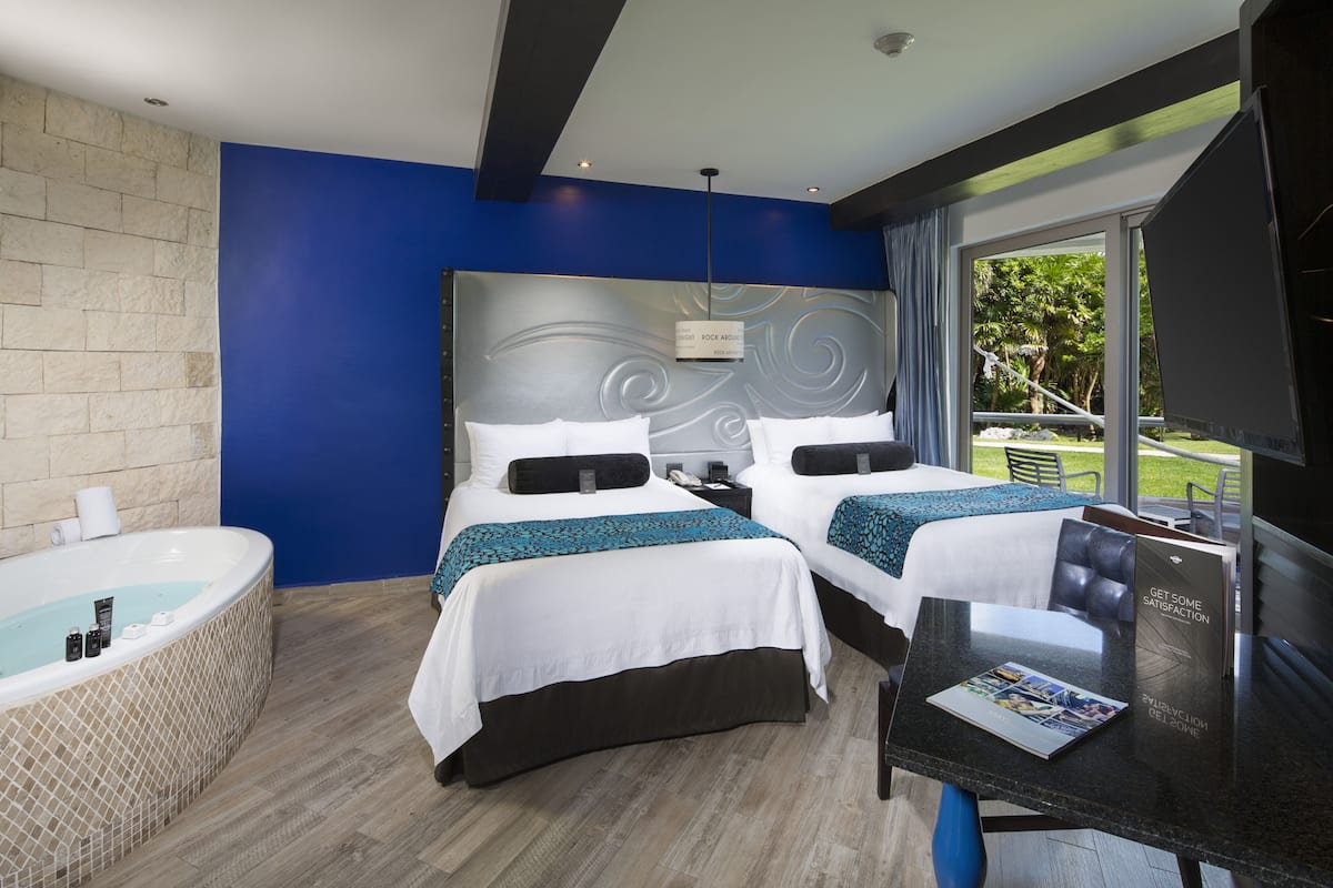 A hotel room with two beds, a large window, a bathtub, and a table with a magazine.