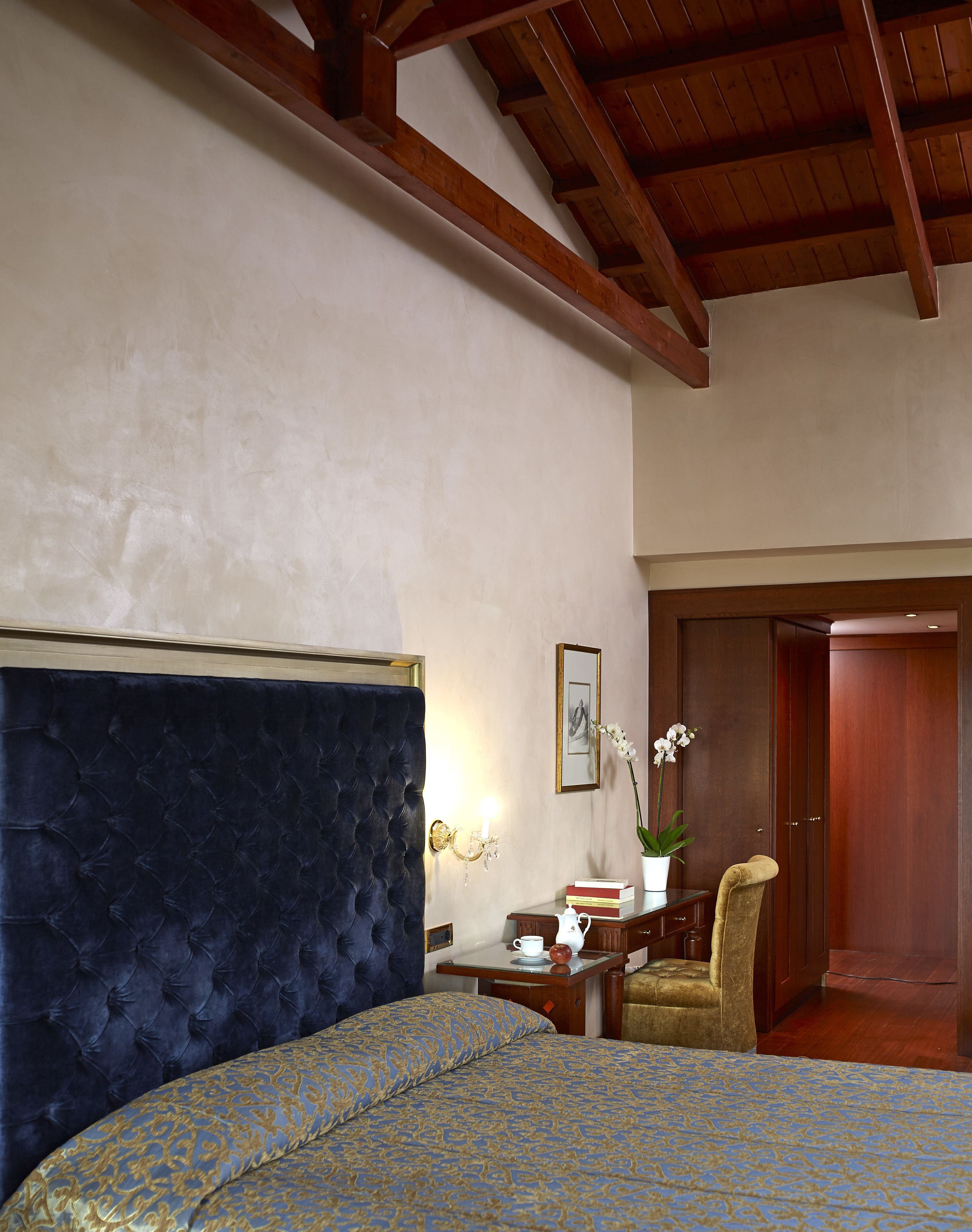A bedroom with a large bed, a wooden ceiling, a nightstand with a lamp, a chair, and a door leading to another room.