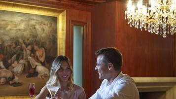 Una pareja sentada a una mesa, con un cuadro en la pared, una araña y una copa de vino.