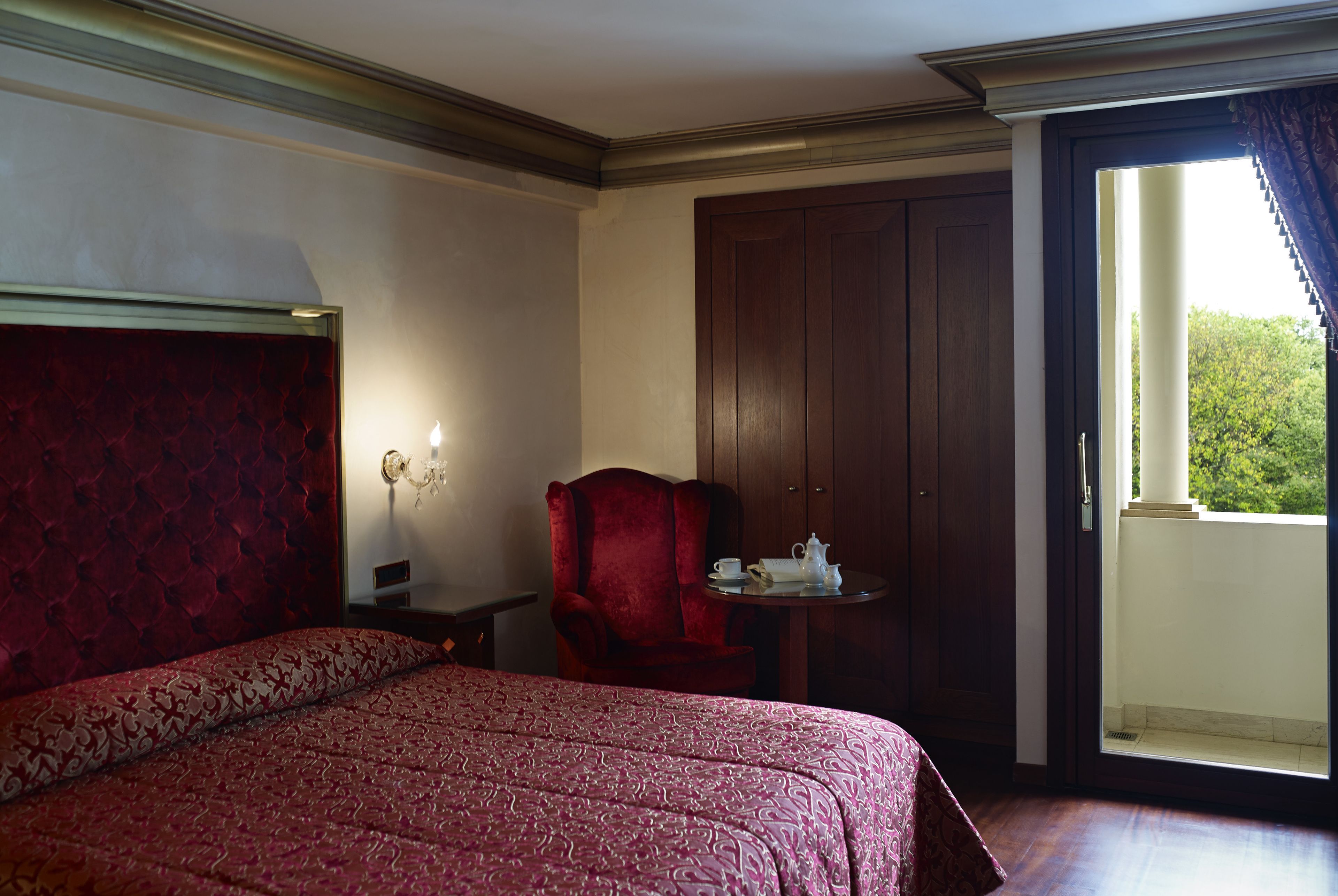 A hotel room with a large bed, a red armchair, a small table with a teapot, and a view of greenery through a window.