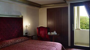 A hotel room with a large bed, a red armchair, a small table with a teapot, and a view of greenery through a window.