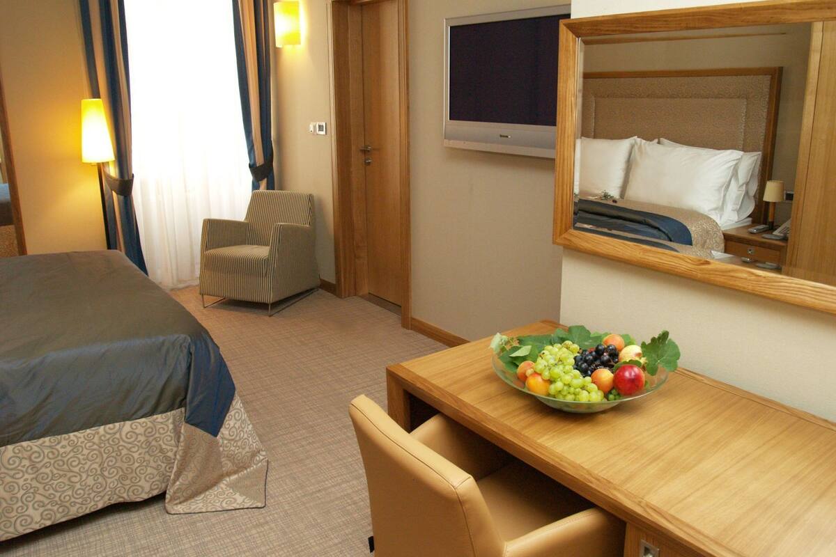 A hotel room with a bed, a desk with a fruit bowl, a chair, a TV, and a door.