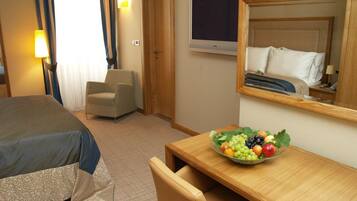 Habitació d'hotel amb un llit, una taula de nit amb un plat de fruita, una cadira, un televisor i una porta.