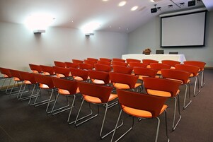 Un salón de conferencias con hileras de sillas anaranjadas, una mesa blanca, una pantalla de proyector y luces en el techo.