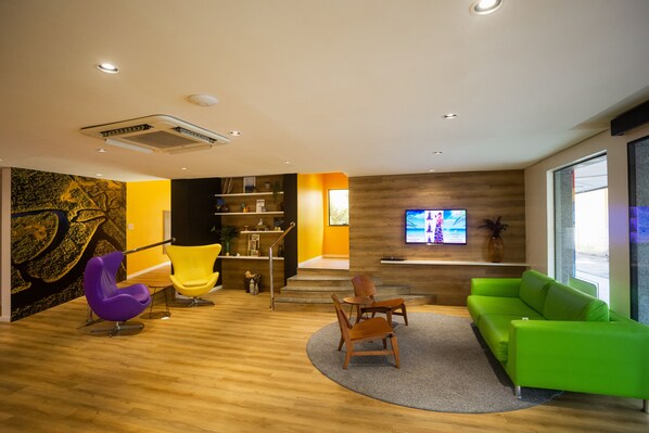 A modern interior with a wooden floor, a green sofa, a purple and a yellow chair, a flat-screen TV, and a staircase leading to a balcony. - ibis Styles Belém Batista Campos (Belém)