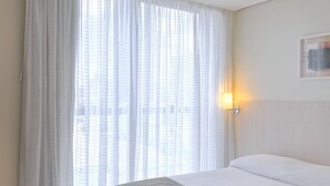 Hotellihuone, jossa on sänky, seinään kiinnitetty valaisin ja ikkuna, jossa on läpikuultavat verhot.