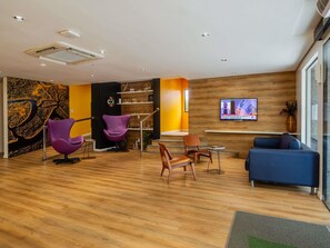 A modern lobby with wooden flooring, purple and brown chairs, a blue sofa, a TV on the wall, and a wooden accent wall. - ibis Styles Belém Batista Campos (Belém)