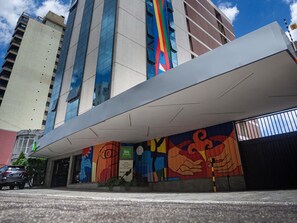 Entrada de un hotel con un mural colorido, un corredor cubierto y un edificio alto al fondo.