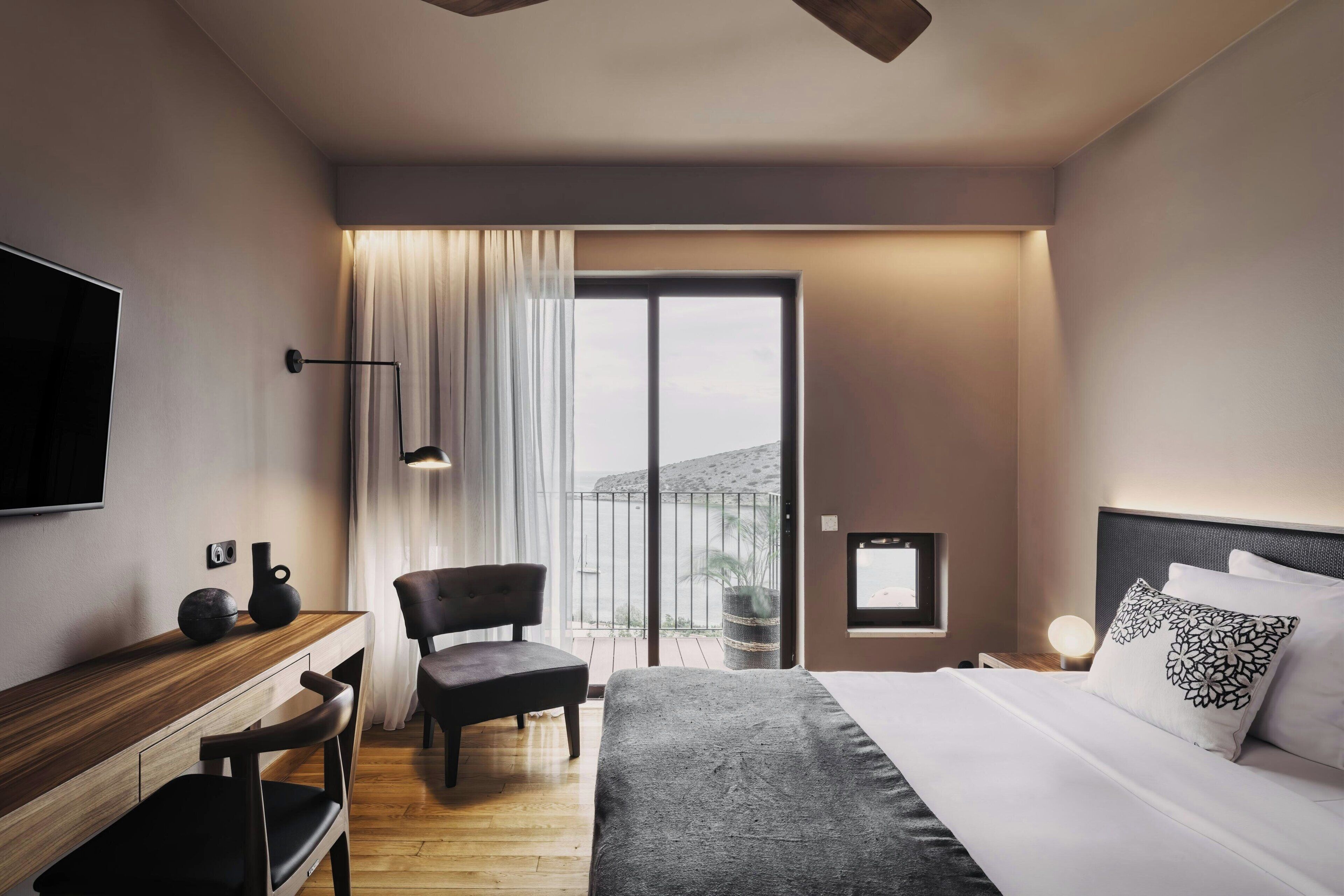 A modern hotel room with a bed, a desk, a chair, a TV, and a balcony with curtains.