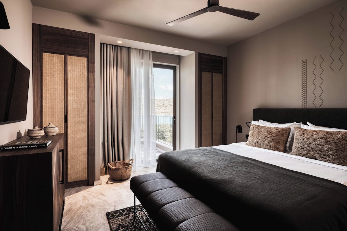 A modern bedroom with a large bed, a ceiling fan, and a view of the outdoors through a window.