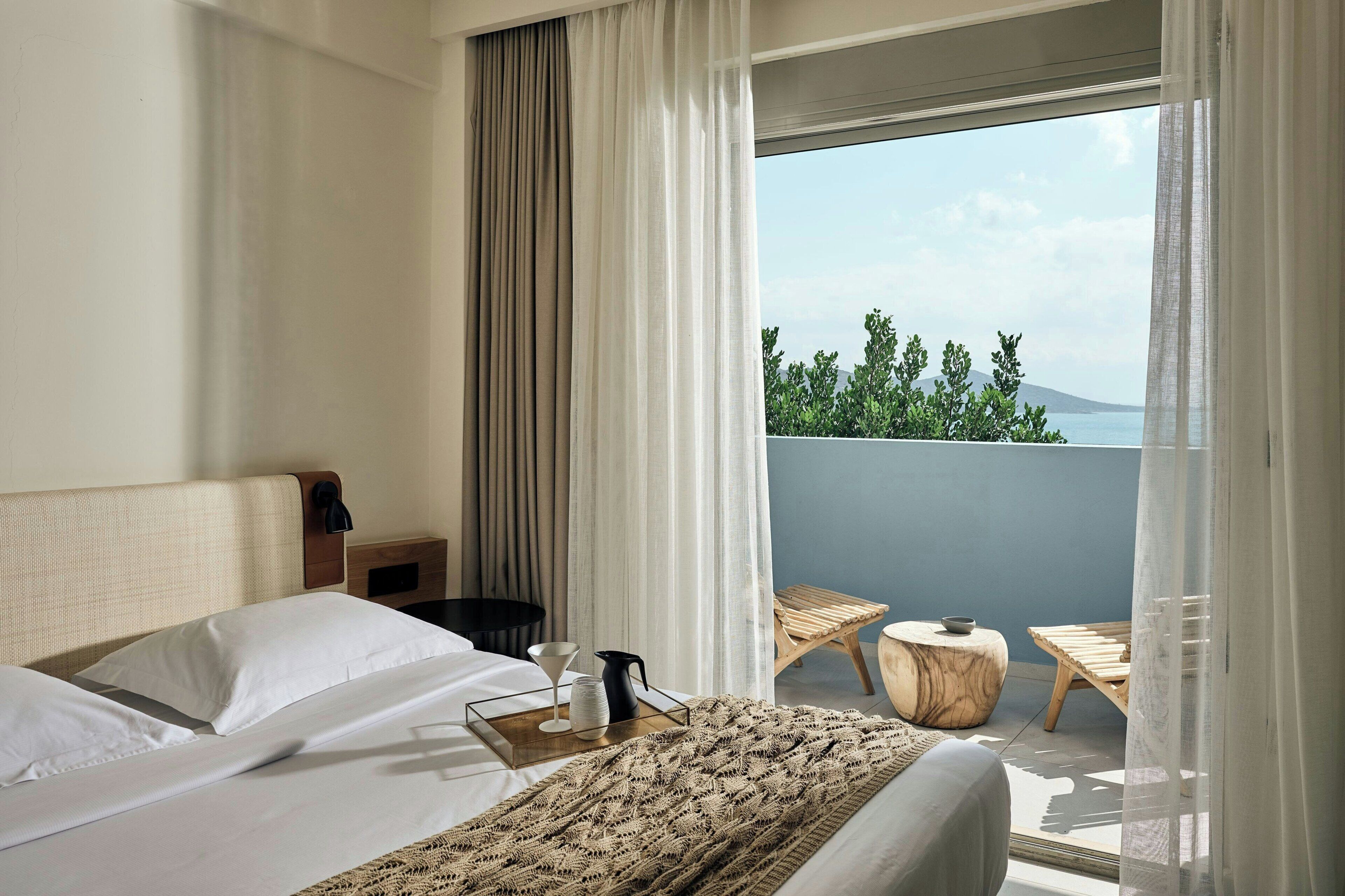 Une chambre d’hôtel comprenant un lit, une table de chevet, une petite table en bois avec un plateau, et donnant sur un balcon aménagé avec du mobilier d’extérieur.