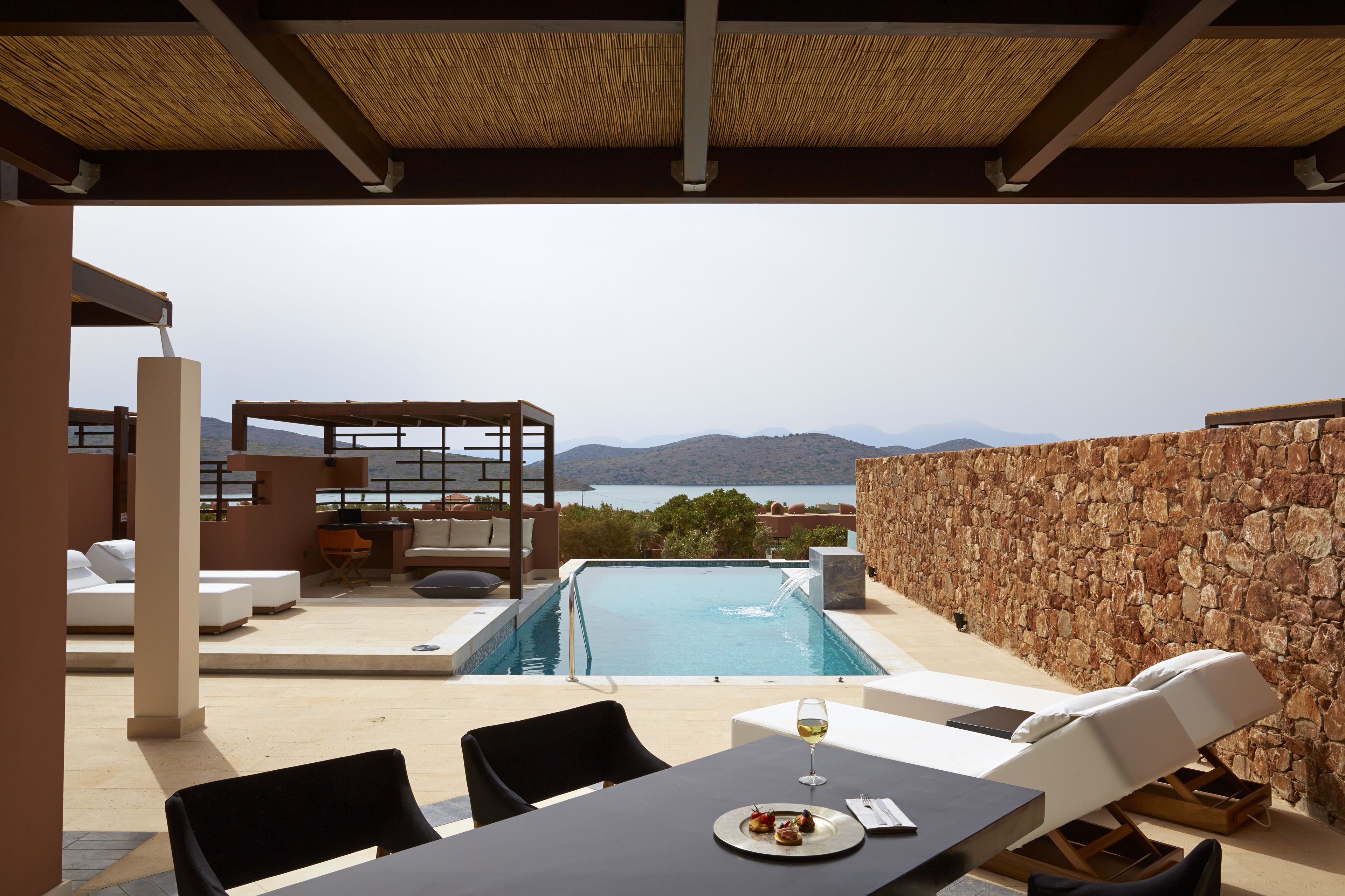 A poolside area with a table, lounge chairs, and a view of the sea and mountains.