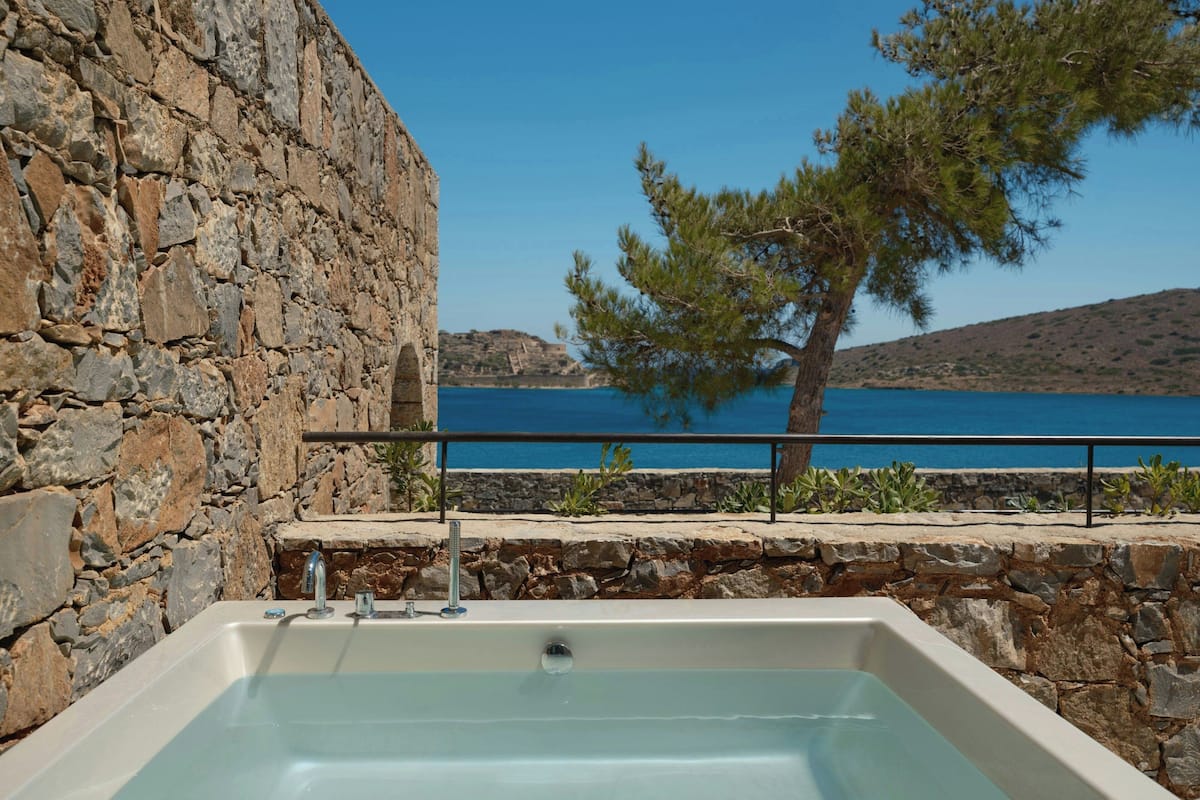 A stone-walled bathroom with a bathtub, a stone bench, and a view of the sea.