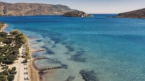 A coastal area with clear blue waters, a sandy beach, and rocky hills in the background. - Domes of Elounda, Autograph Collection (Agios Nikolaos)