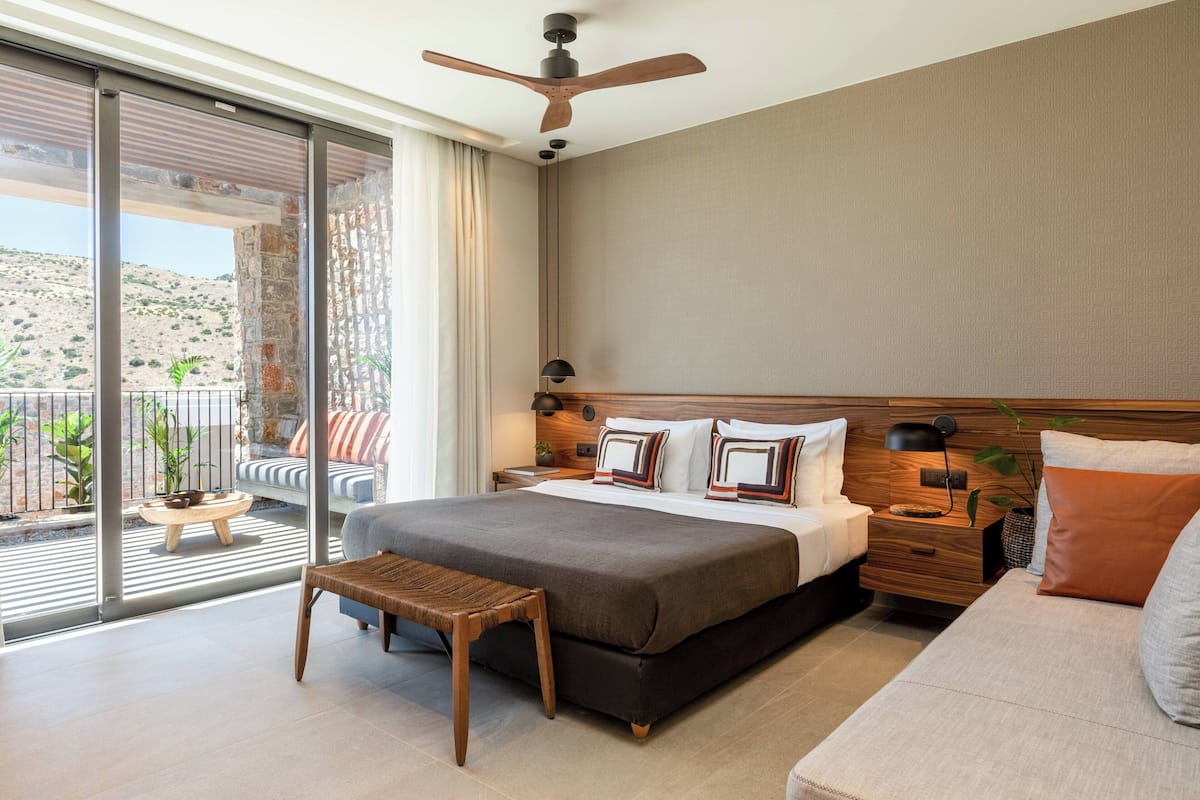 A modern hotel room with a large bed, a wooden headboard, a nightstand, a bedside table, a bench, and a view of the outdoors through a sliding glass door.