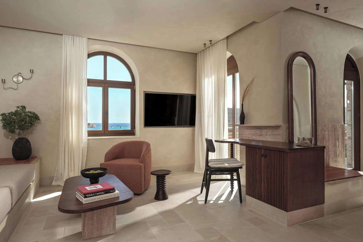 A modern living room with a large window, a flat-screen TV, a sofa, a coffee table with books, a chair, and a wooden cabinet.