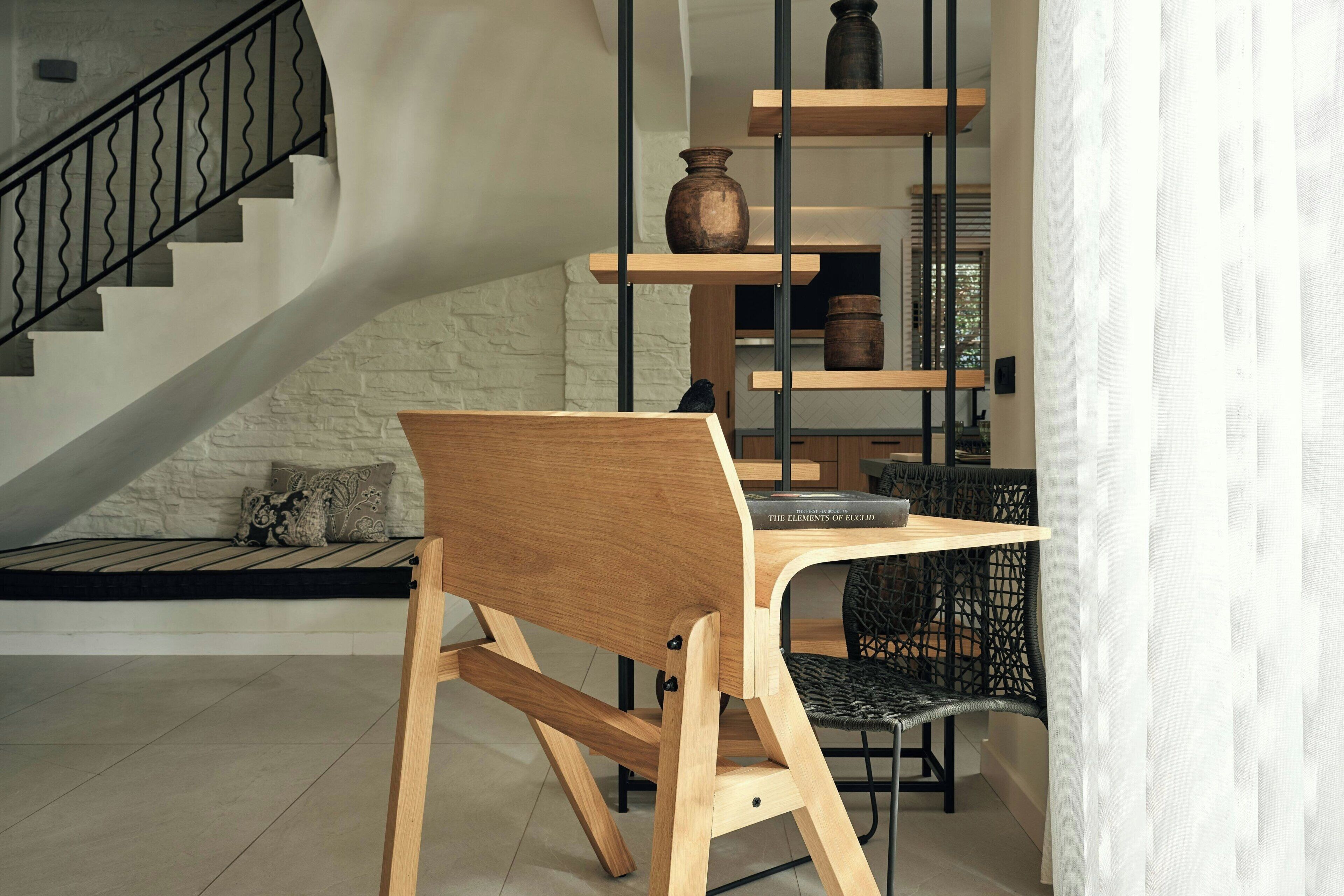 Ein Essbereich mit einem Holztisch und Stuhl, ein schwarzer Stuhl mit Sitzgeflecht, eine Treppe mit schwarzer Geländer und Regale mit Keramik und Büchern.