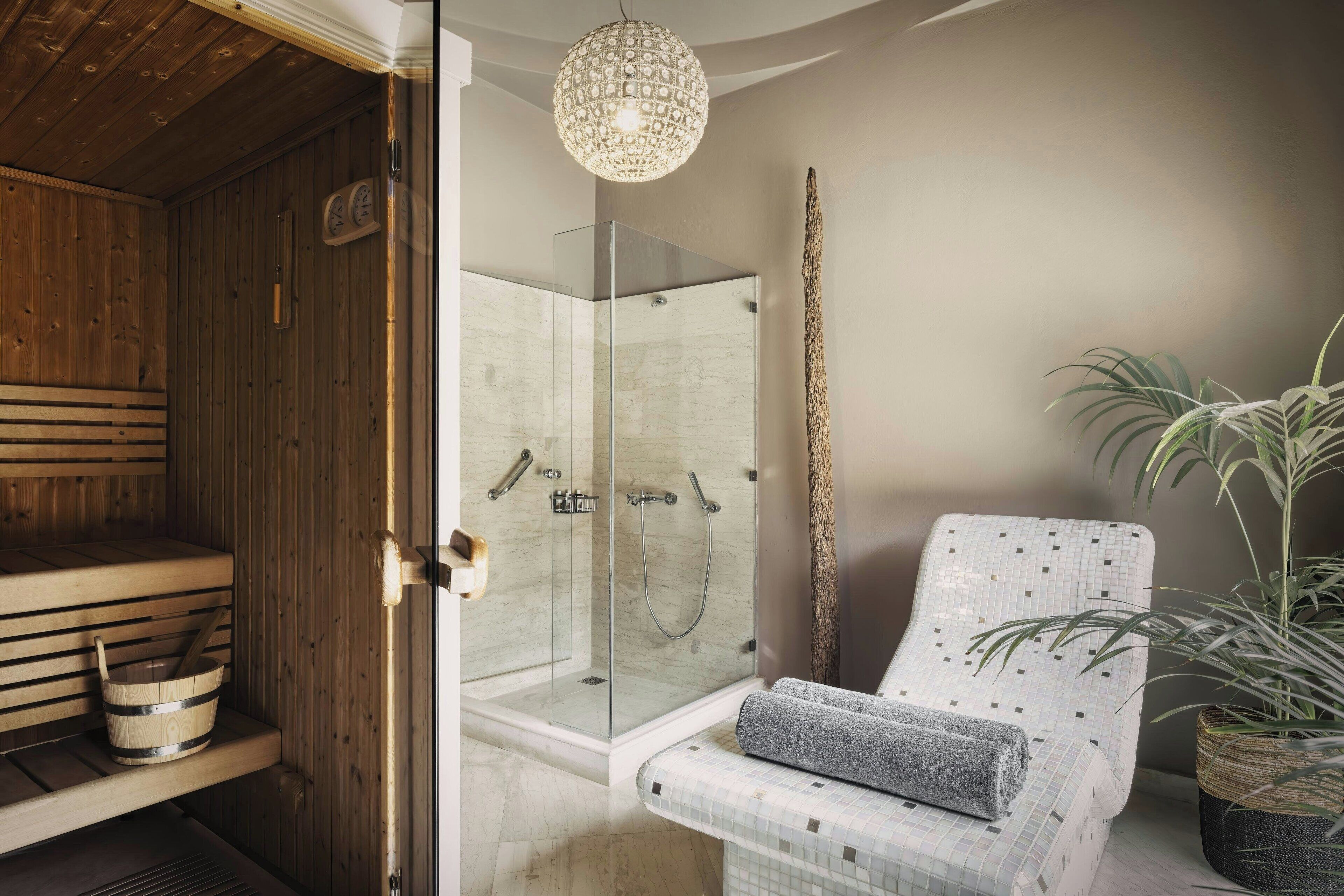 A modern bathroom with a glass-enclosed shower, wooden sauna benches, a seating area with a patterned chair, and a potted plant.