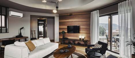 A modern living room with a white sofa, a round wooden coffee table, and a grey armchair. There is a flat-screen TV mounted on the wall, a dining table with chairs, and a large window with curtains.