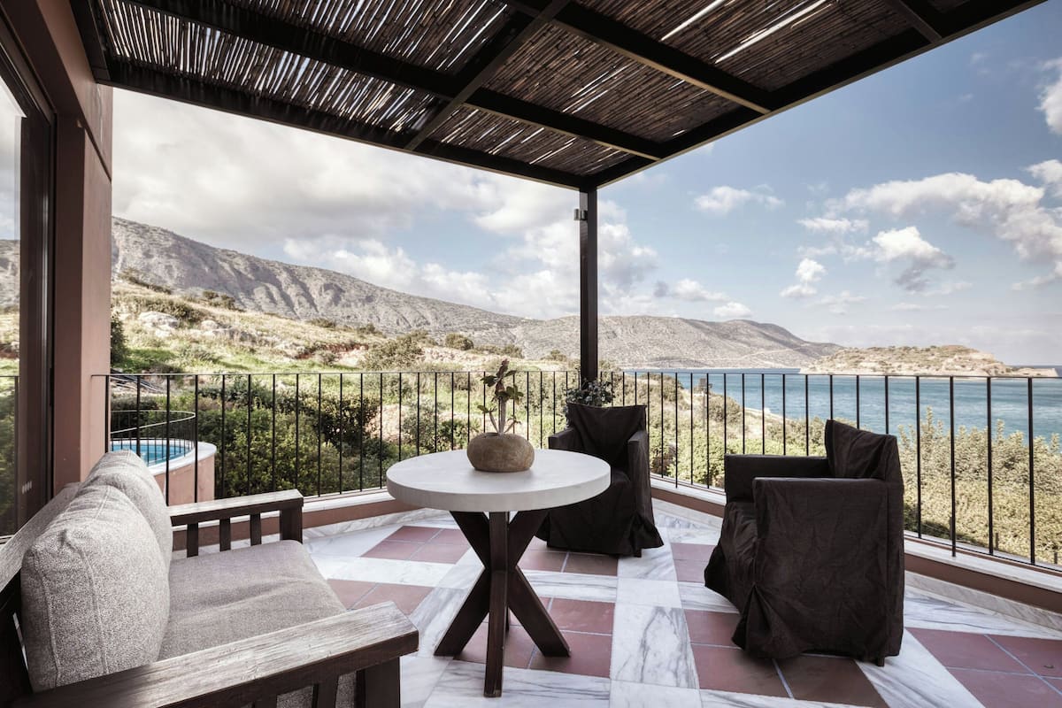 A balcony with a round table, seating, and a view of the sea and mountains.