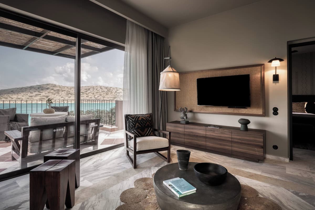 A modern living room with a large TV, a comfortable sofa, a dining area, and a view of the sea.