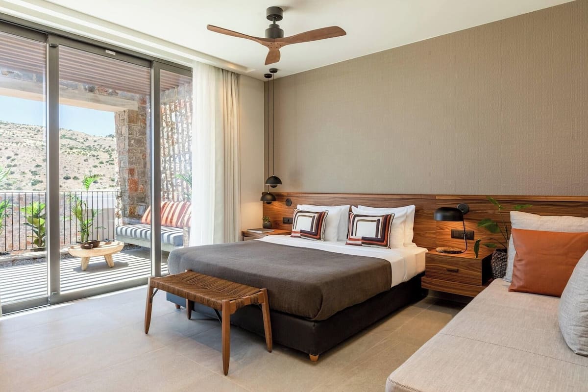 Una habitación de hotel moderna con una cama grande, un cabecero de madera, una mesita de noche y vistas a un balcón con mobiliario exterior.