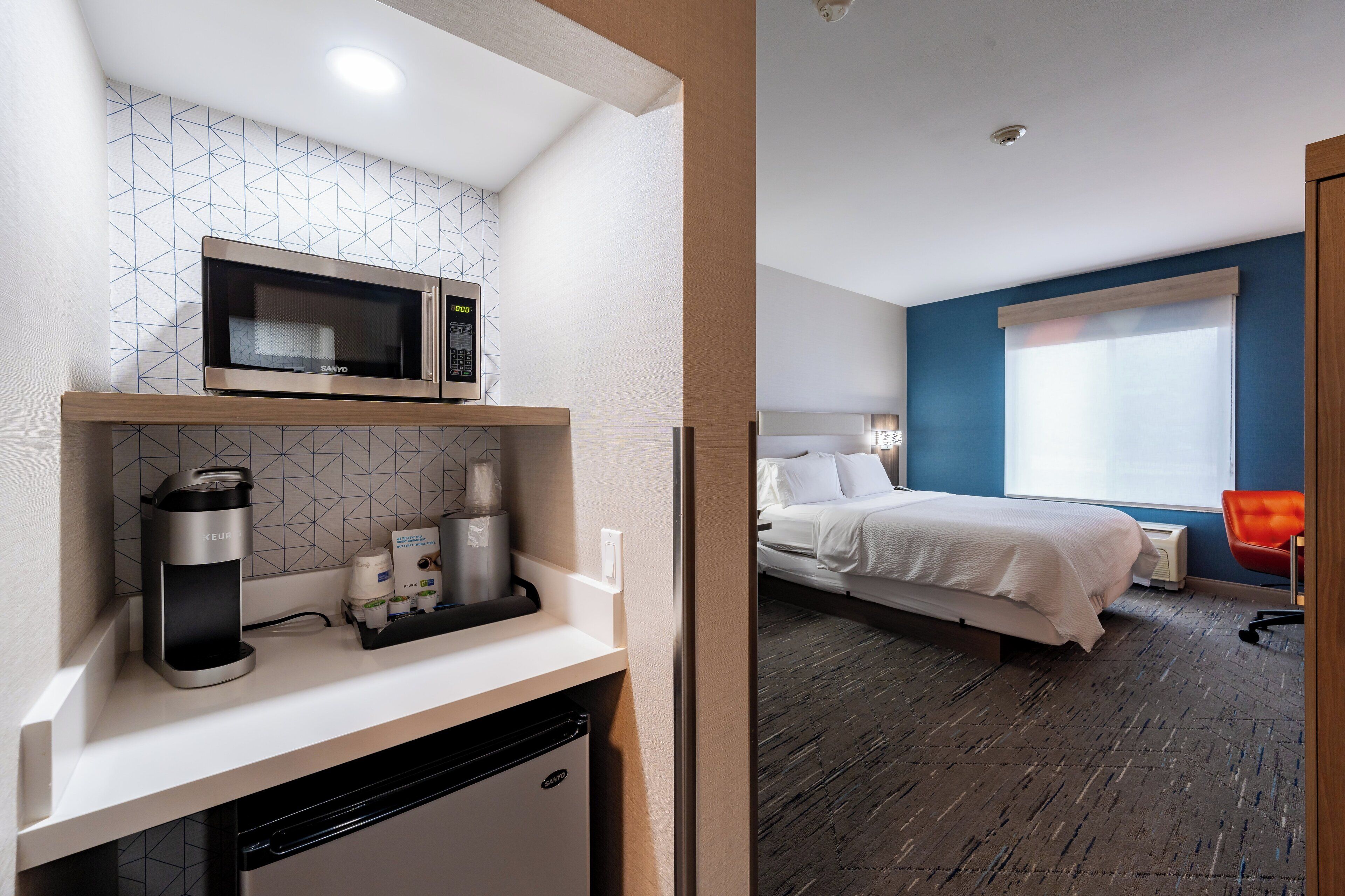 Une petite cuisine équipée d’un four à micro-ondes, d’une machine à café et d’un petit réfrigérateur. À côté de la cuisine se trouve une chambre avec un lit, une table de chevet et une fenêtre donnant sur un paysage agréable.