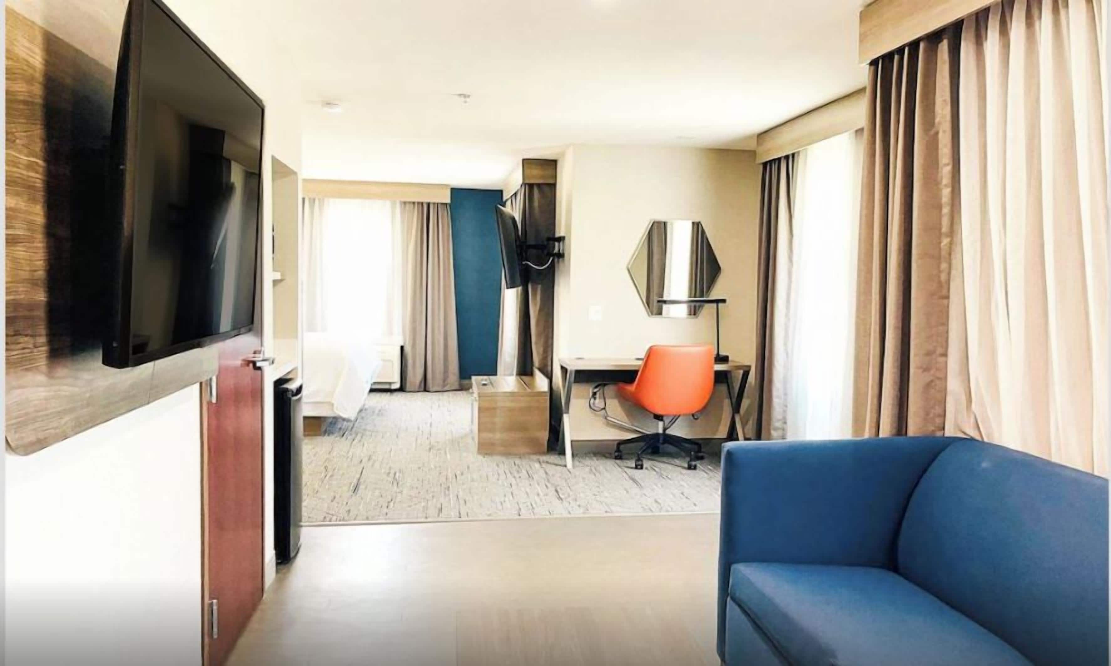 A hotel room with a flat-screen TV, a blue sofa, a desk with an orange chair, a bed with white linens, and a large window with curtains.