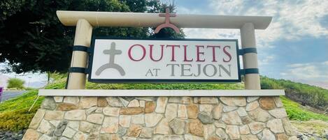 A sign for 'OUTLETS AT TEJON' with a cross symbol, set against a stone wall and surrounded by rocks.