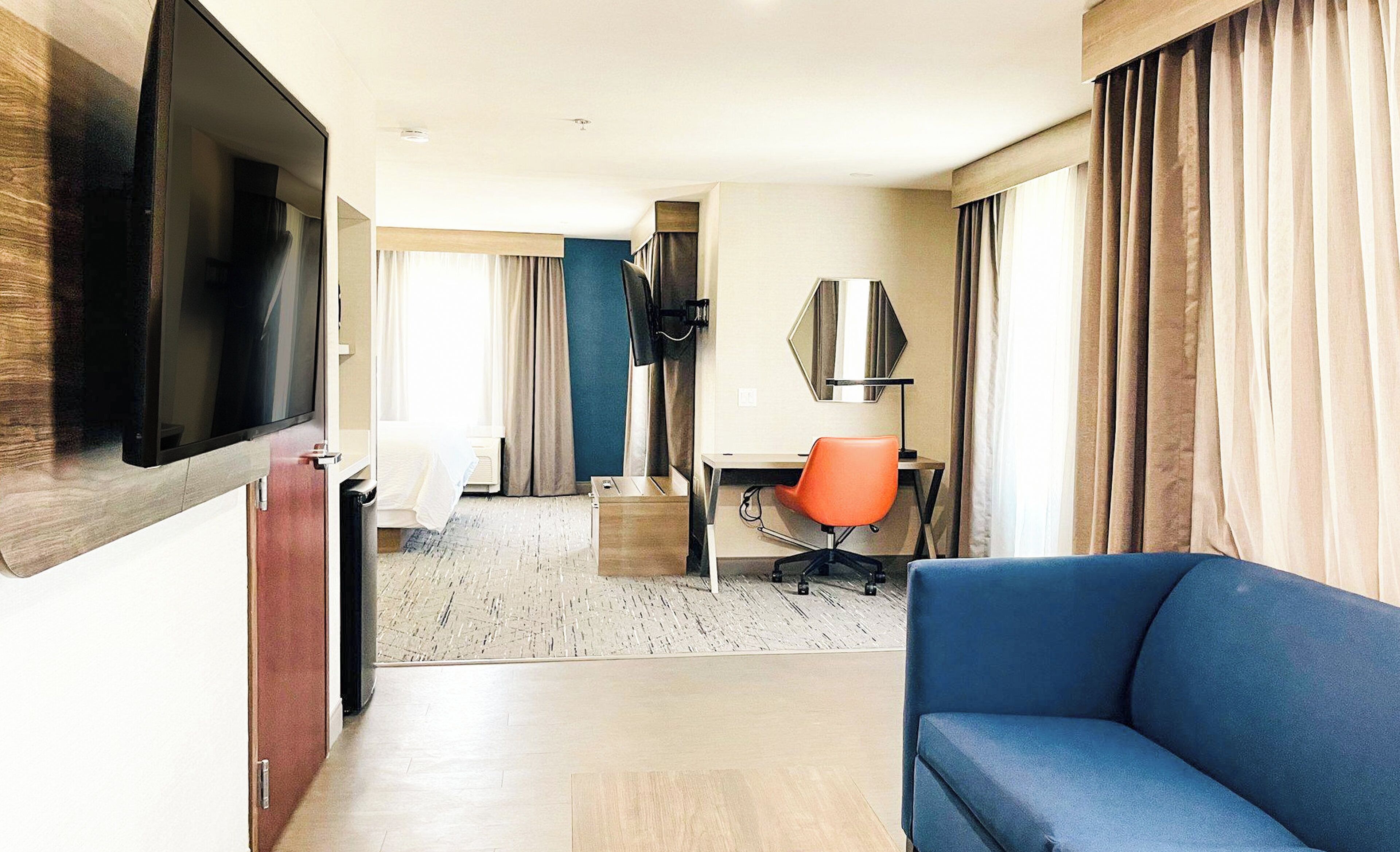 Ein Hotelzimmer mit Fernseher, einem blauen Sofa, einem orangefarbenen Bürostuhl, einem Schreibtisch und einem großen Fenster mit Vorhängen.