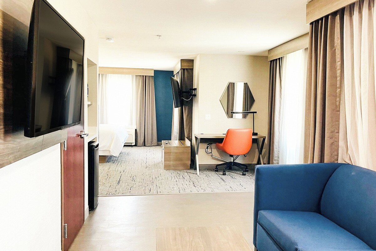 A hotel room with a TV, a blue sofa, an orange office chair, a desk, and a large window with curtains.
