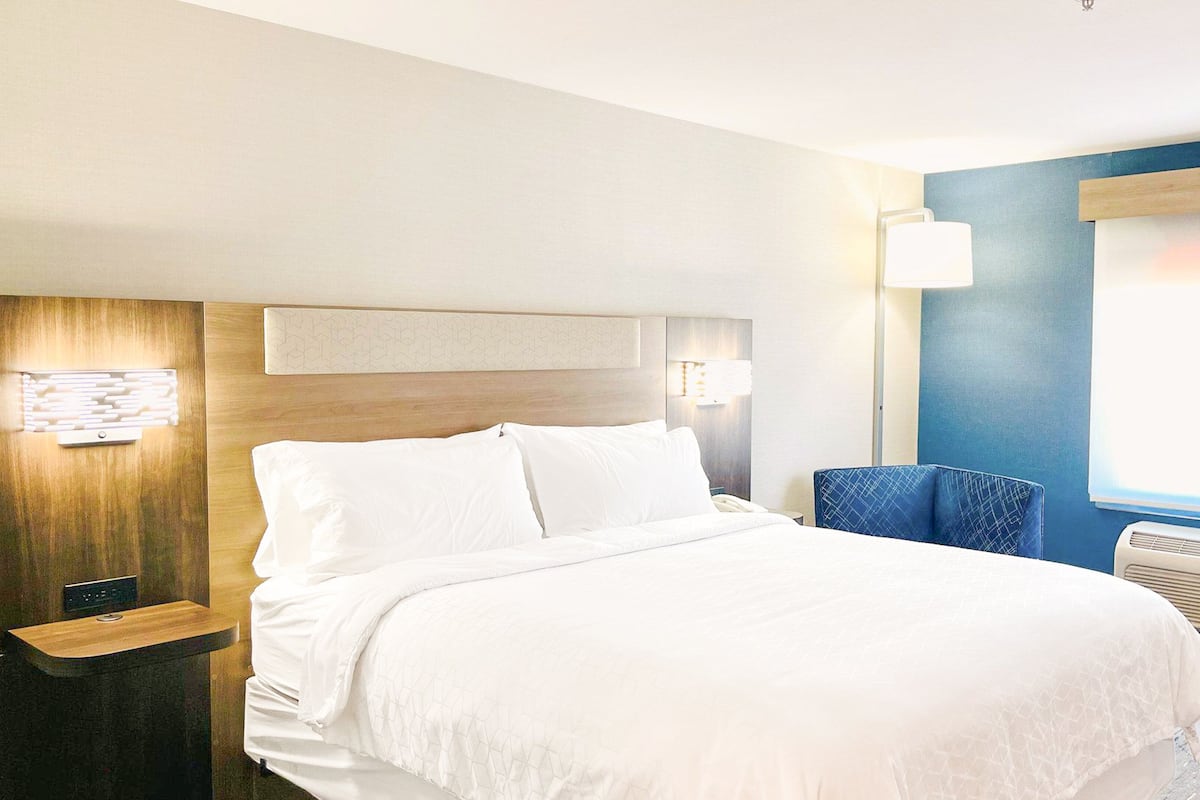 A hotel room with a bed, bedside table, lamp, chair, and air conditioning unit.
