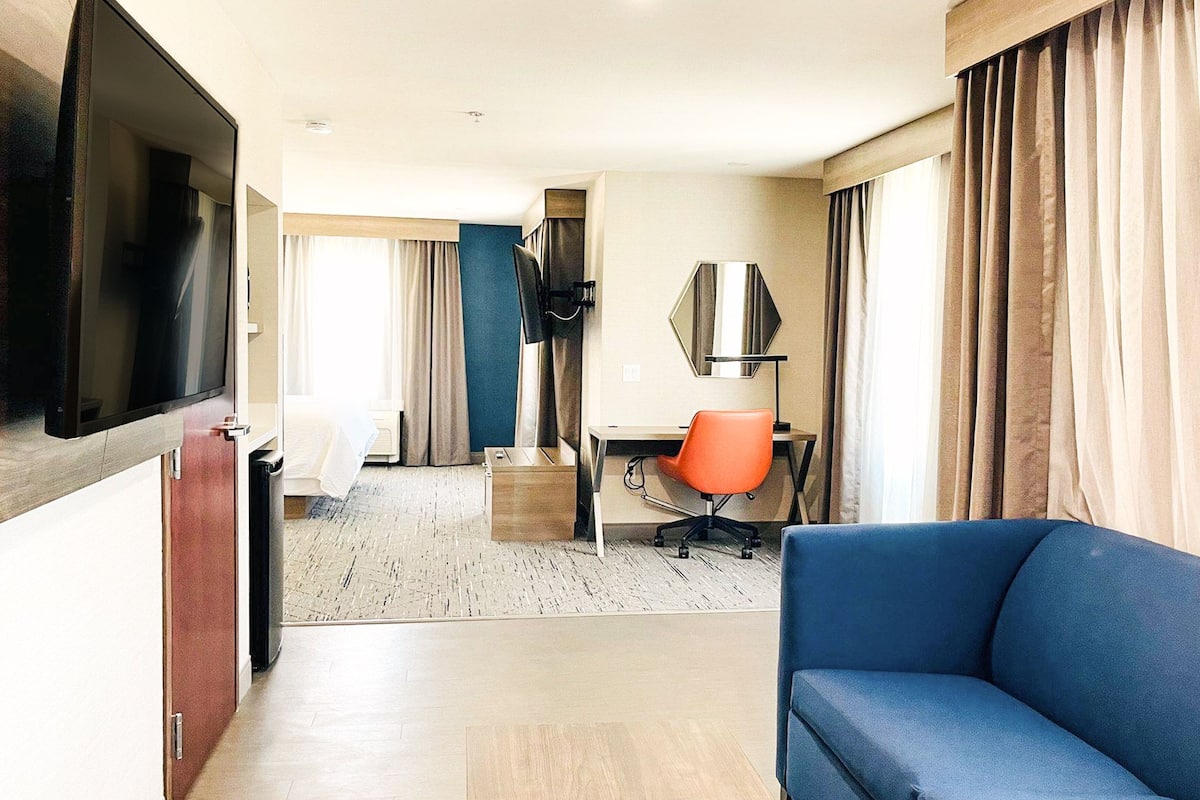 A hotel room with a TV, a blue sofa, an orange office chair, a desk, and a large window with curtains.
