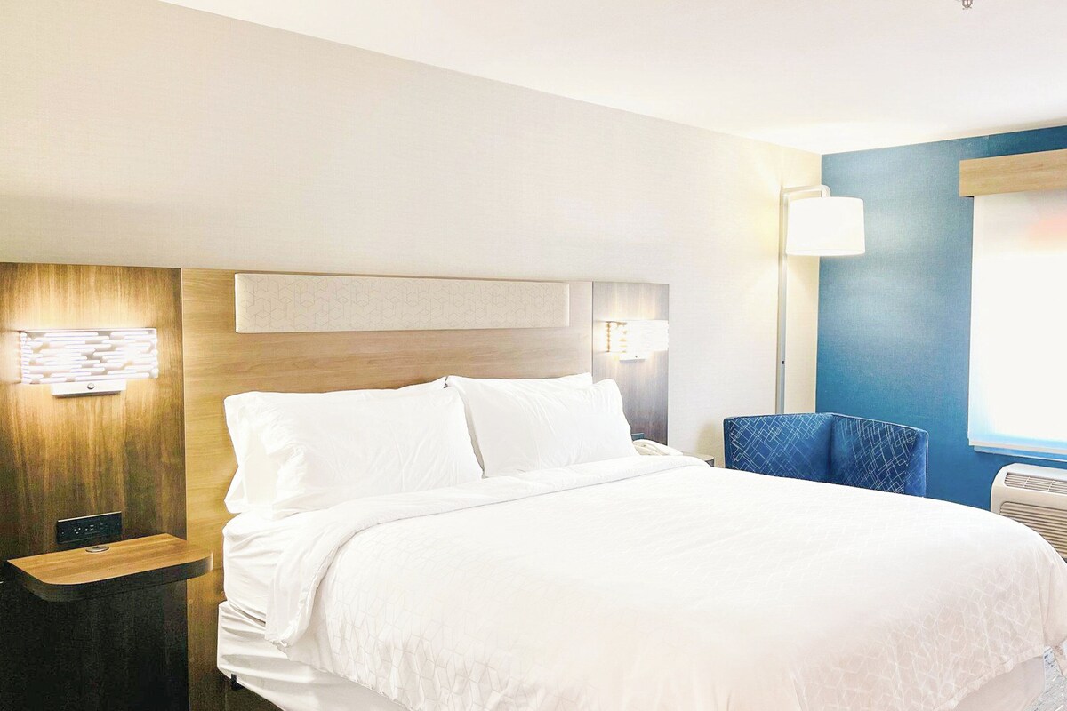 A hotel room with a bed, bedside table, lamp, chair, and air conditioning unit.