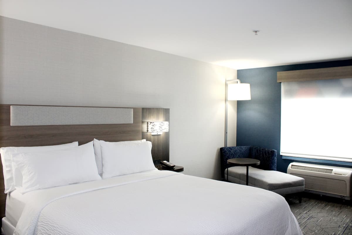 A hotel room with a large bed, a desk, a chair, a lamp, and a window with blinds.