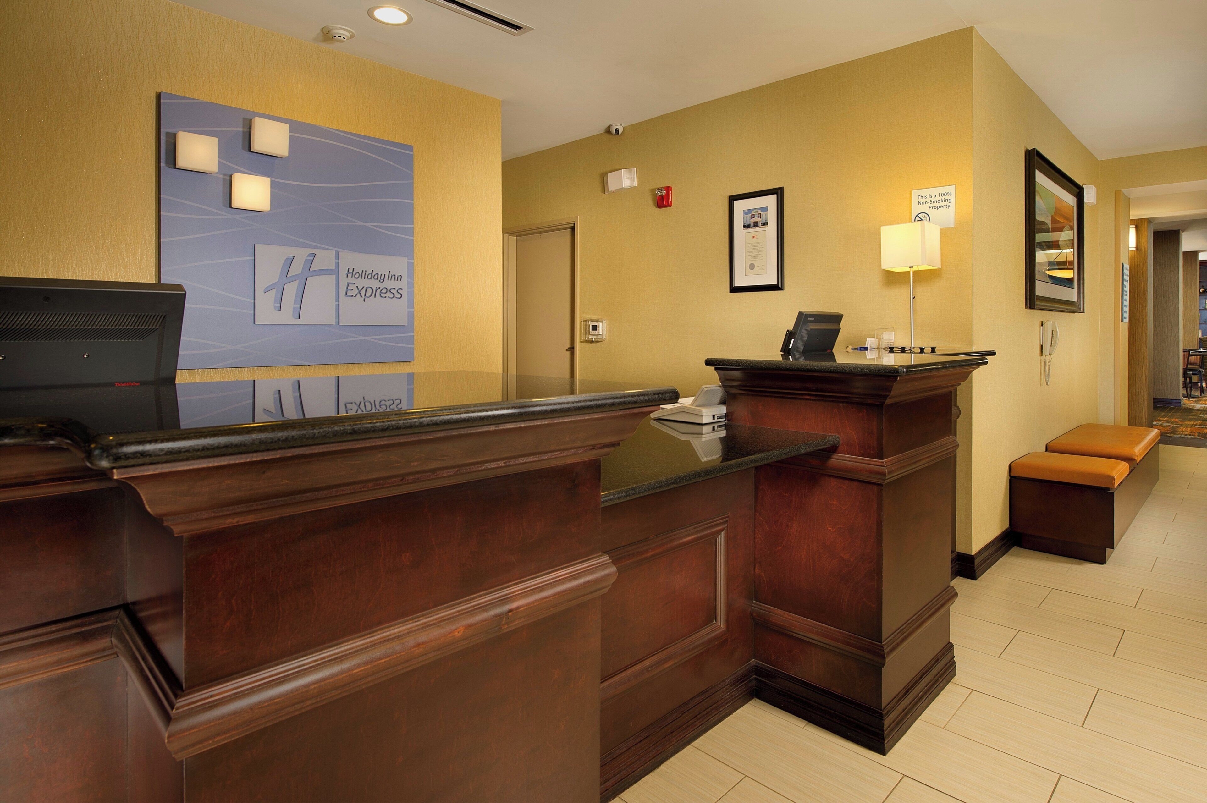 A hotel reception desk with a wooden counter, a Holiday Inn Express sign, a security camera, and a framed certificate on the wall.