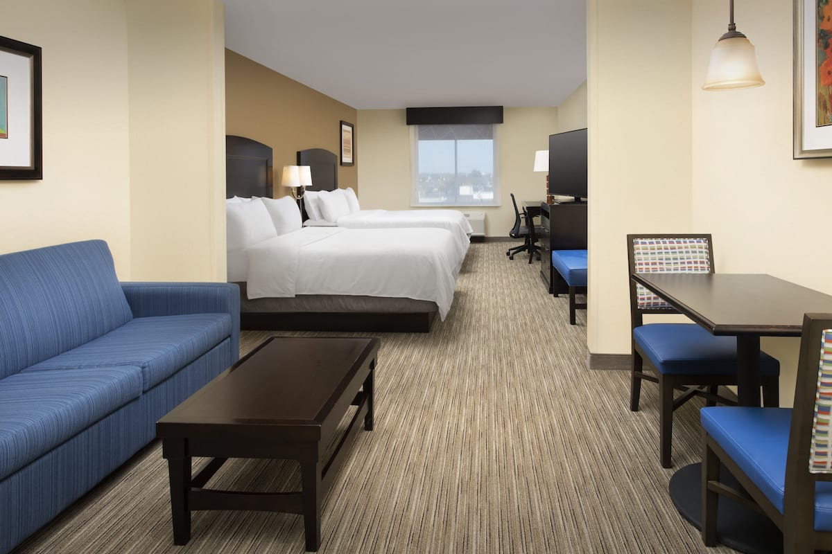 A hotel room with a blue sofa, two single beds, a desk with a chair, and a view of the city through the window.
