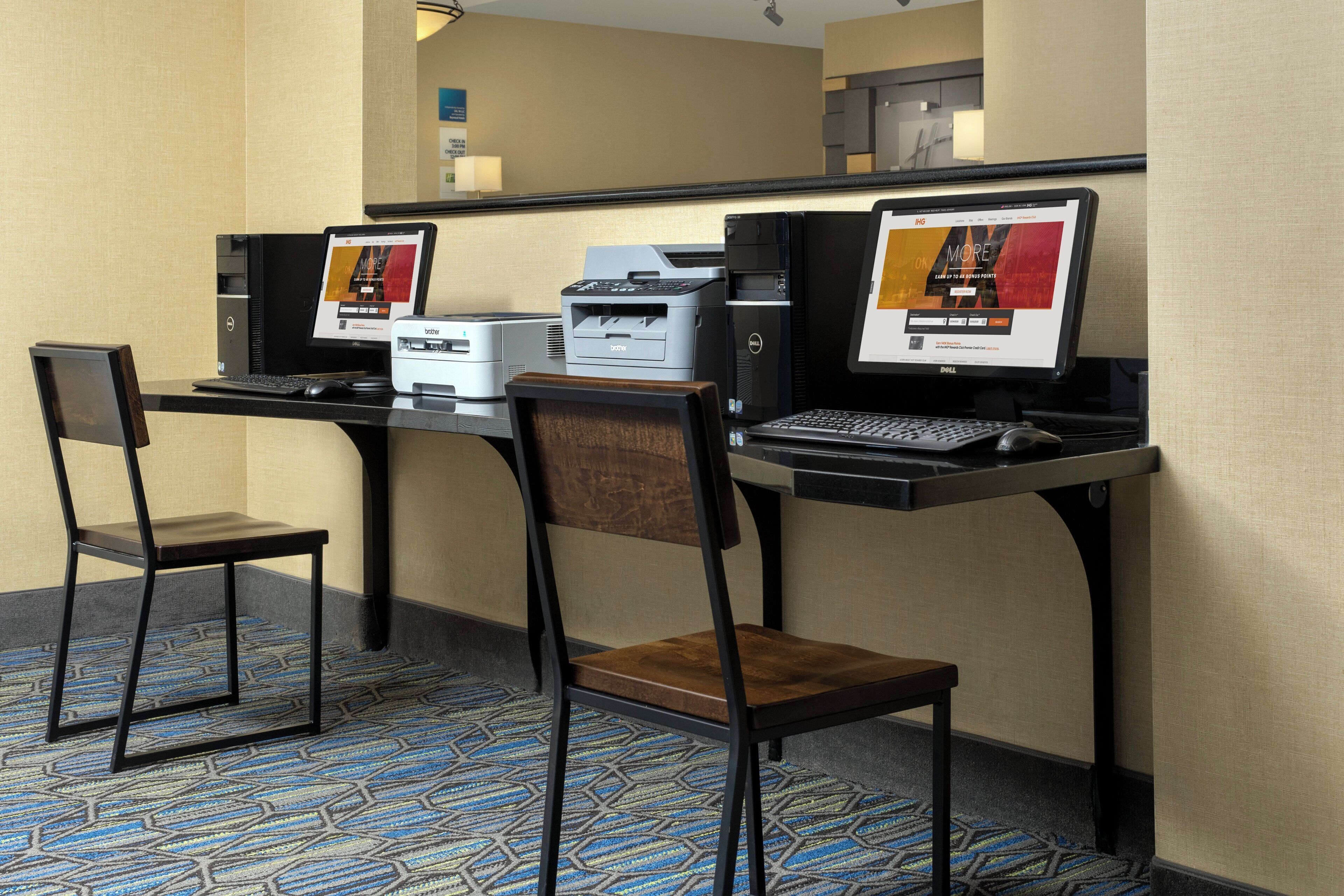 A hotel room with a desk, two chairs, a desktop computer, a printer, and a laptop.