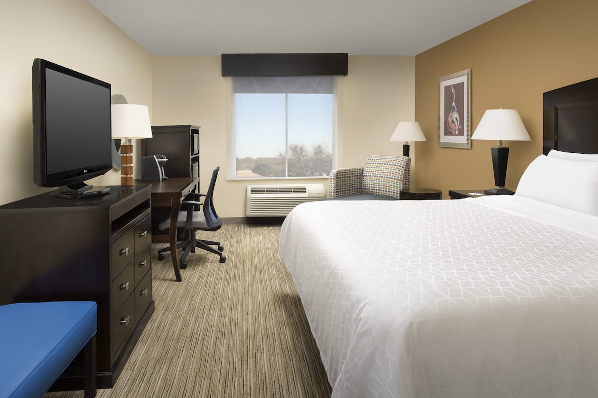 A hotel room with a large bed, a desk with a computer, a chair, a television, and a window with a view of trees.