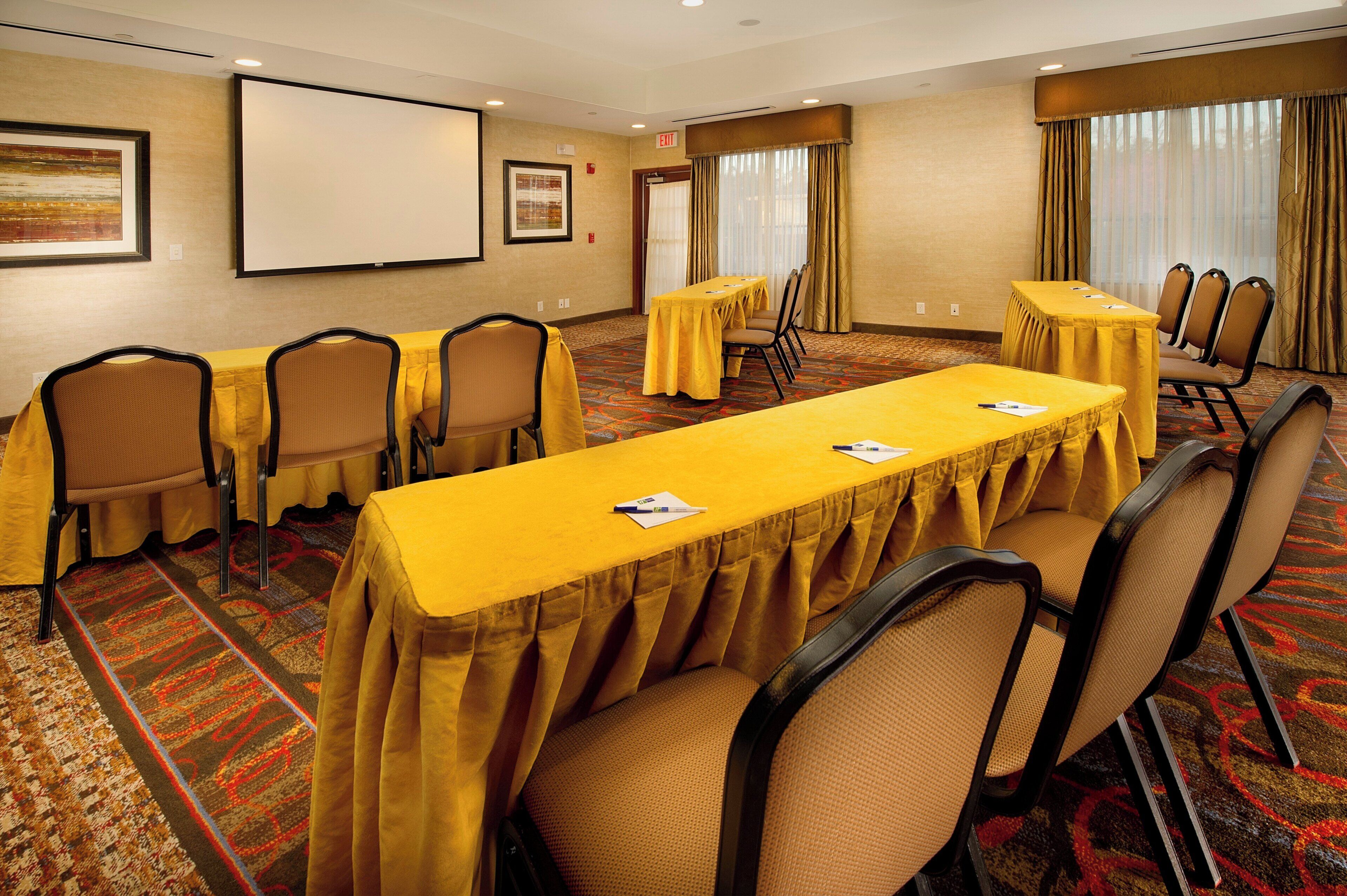 A conference room with a long table, multiple chairs, a projector screen, and a carpeted floor.