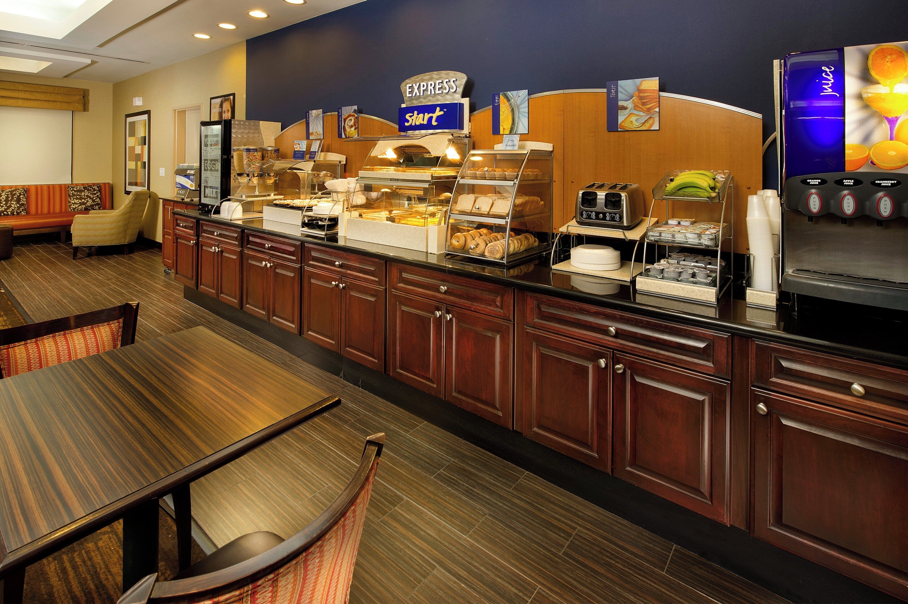 Un área de desayuno de hotel con una barra de buffet, mesas y sillas, y un dispensador de jugo.